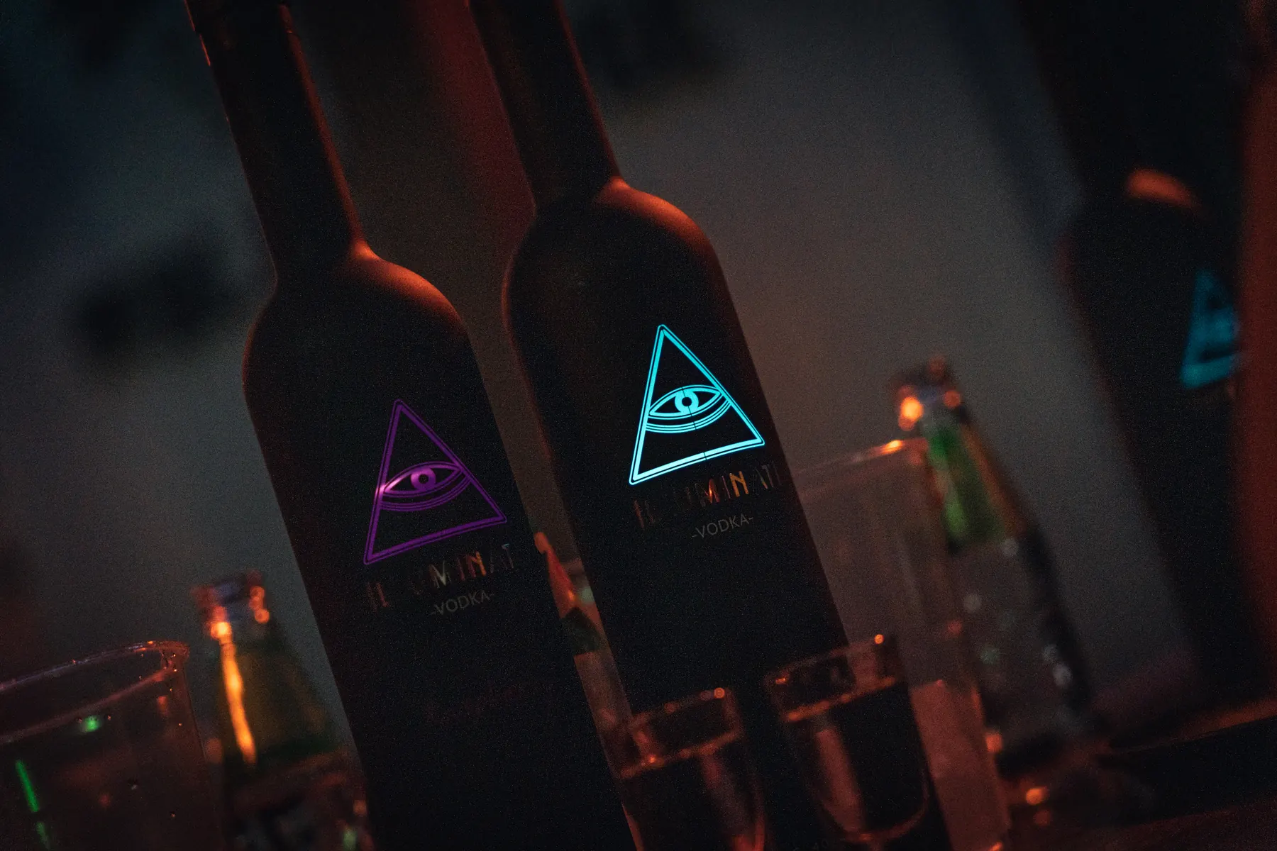 Two Illuminati Vodka bottles in low light, the Raspberry eye glowing pink and the Original eye glowing blue